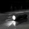 A black flashlight with green accents is lying on a wet, dark surface, emitting a bright light. The background is dark and out of focus, suggesting an outdoor night setting. Produktbild-Vorschau