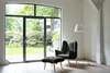 A modern living room with a large window overlooking a garden, featuring a dark gray armchair and ottoman, and a white floor lamp. Produktbild-Vorschau