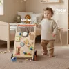A baby is standing next to a wooden walker in a nursery. Produktbild-Vorschau