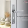 A close-up of a silver door handle and escutcheon plate mounted on a white door, with a blurred background of a room interior. Produktbild-Vorschau