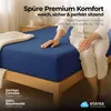 A person sitting on a bed with a blue fitted sheet. Produktbild-Vorschau