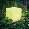 A glowing yellow cube-shaped solar light sits in a garden surrounded by plants and grass. Produktbild-Vorschau