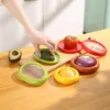 A hand is shown placing an avocado half into a green food storage container on a wooden cutting board. Other food items and containers are also visible. Produktbild-Vorschau
