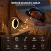 A woman sleeping in bed next to a bedside table with a white noise machine and alarm clock. Produktbild-Vorschau
