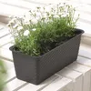 A dark brown planter with white flowers sits on a white wooden surface. Produktbild-Vorschau
