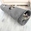 Two kittens playing in a gray fabric tunnel on a wooden floor, with a couch in the background. Produktbild-Vorschau
