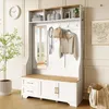 A white and wood-tone hall tree with a mirror, bench, drawers, and cabinets, set against a white paneled wall with decorative molding. A plant is in the foreground, and a framed artwork hangs on the wall. Produktbild-Vorschau