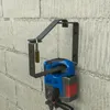 A black metal wall mount holding a blue and red power tool, secured with a brass padlock, mounted on a concrete block wall. Produktbild-Vorschau