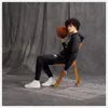 A young man sits on a wooden chair holding a basketball, wearing a black hoodie and black sweatpants with white sneakers. Produktbild-Vorschau