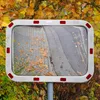 A convex traffic mirror with red and white reflectors on its frame, mounted on a pole, with autumn foliage in the background. Podgląd zdjęcia produktu
