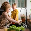 Two children are in a kitchen, with the girl washing an apple under a faucet and the boy watching. Produktbild-Vorschau