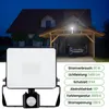A floodlight with a motion sensor is shown in the foreground, with a house and carport in the background at dusk. Náhled obrázku produktu