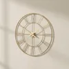 A gold-colored wall clock with Roman numerals is mounted on a light gray wall, casting a shadow. Voir les images du produit