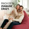 A couple is embracing and sitting on a large red beanbag chair in a living room. Voir les images du produit