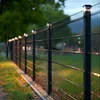 A black metal fence with solar lights on top, illuminated by warm outdoor lighting, with grass and fallen leaves in the foreground. Voir les images du produit