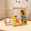 Two children playing on a wooden indoor climbing frame with a slide, climbing wall, rope net, and basketball hoop. Podgląd zdjęcia produktu