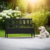 A black wooden garden bench with a storage compartment is placed on a paved patio next to a lawn and a bush with white flowers. A golden retriever dog is lying on the patio to the right of the bench. Anteprima dell’immagine prodotto