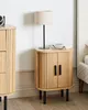 A wooden nightstand with two doors and a lamp on top, placed next to a bed. Náhľad obrázku produktu