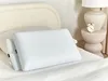 Two white pillows are placed on a bed with a textured headboard in the background. A small alarm clock and a lamp are visible on a side table. Náhľad obrázku produktu