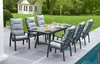 An outdoor dining set with chairs and a table on a patio, with a garden and a small building in the background. Produktbild-Vorschau