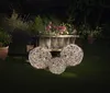 Three illuminated spherical decorative lights are placed on a lawn in a garden at night, with a flower-filled wooden cart in the background. Produktbild-Vorschau