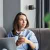 A woman is sitting on a couch, holding a phone and looking away from the camera. A laptop is on her lap. A wall charger is plugged into the wall behind her, charging multiple devices. Produktbild-Vorschau