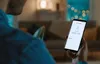 A person is holding a smartphone in a dimly lit room, with a couch and decorative lights in the background. Náhled obrázku produktu