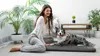 A woman is sitting on the floor next to her dog, who is lying on a dog bed. The scene is indoors, with plants and furniture in the background. Náhľad obrázku produktu