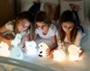 Three young girls lying on a bed, looking at several cute, glowing silicone night lights shaped like animals. Produktbild-Vorschau