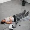 A woman is lying on a yoga mat with her legs elevated and encased in compression boots, connected to a device. A remote control is nearby. Produktbild-Vorschau