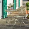 A metal bistro set with a table and chair on a wooden deck, with green shutters and potted plants in the background. Náhled obrázku produktu