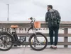 A man with a backpack stands next to an electric bicycle on a wooden pier. Voir les images du produit