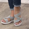 Graue Stoffsandalen mit geknotetem Riemendesign an den Füßen einer Person, die Jeans mit Aufschlag trägt, auf Holzboden stehend Anteprima dell’immagine prodotto