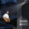 A delivery person carrying a box approaches a house at night, illuminated by a security camera's spotlight. Produktbild-Vorschau