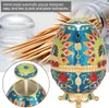 A decorative egg-shaped toothpick holder with peacock and rose designs, shown with a pile of toothpicks in the background and a close-up inset of the lid mechanism. Produktbild-Vorschau