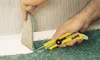 A person is using a utility knife to cut carpet next to a wall. Produktbild-Vorschau