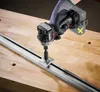 A gloved hand is using a cordless impact driver to attach a metal bracket to a long metal rail on a wooden workbench. Produktbild-Vorschau