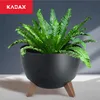 A black planter with wooden legs holding a green plant, set against a dark gray background. Náhled obrázku produktu