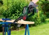 A person is using a cordless chainsaw to cut a piece of wood outdoors. Produktbild-Vorschau
