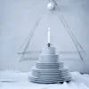 A stack of white plates arranged in a pyramid shape, topped with a lit candle in a silver holder. A Christmas tree shape is drawn on the wall behind, with a white ornament at the top. The scene is set on a white tablecloth. Produktbild-Vorschau
