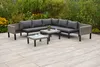 An outdoor seating area with a sectional sofa and coffee tables on a paved patio, surrounded by greenery and flowers. Produktbild-Vorschau