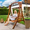 A woman is sitting on a wooden swing chair in a garden, reading a book. Náhled obrázku produktu