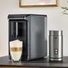 A coffee machine and milk frother are displayed on a dark countertop, with a glass of layered coffee in front of the machine. Produktbild-Vorschau