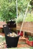 A black garden cart filled with leaves and debris is shown outdoors next to raised garden beds. Náhled obrázku produktu