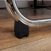 A close-up view of a black protective foot attached to a chrome metal tube, resting on a wooden floor. Voir les images du produit