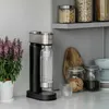 A soda maker sits on a kitchen counter next to a potted plant and jars of food. Podgląd zdjęcia produktu