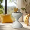 A stylish side table with a unique hourglass shape, placed in a well-lit living room setting next to a window with a view of nature. A yellow pillow and a yellow throw blanket add pops of color to the scene. Anteprima dell’immagine prodotto