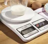 A digital kitchen scale with a cup of liquid on it, placed on a wooden surface. Eggs and a plate are visible in the background. Voir les images du produit