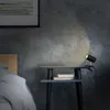 A black clip-on lamp is attached to a small bedside table, illuminating a stack of books and the textured wall behind it. Náhled obrázku produktu