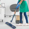 Blauer Flachwischer mit grauem Mikrofaserpad, benutzt von einer Person mit blauen Handschuhen und grüner Schürze, um einen weißen Boden zu reinigen Produktbild-Vorschau